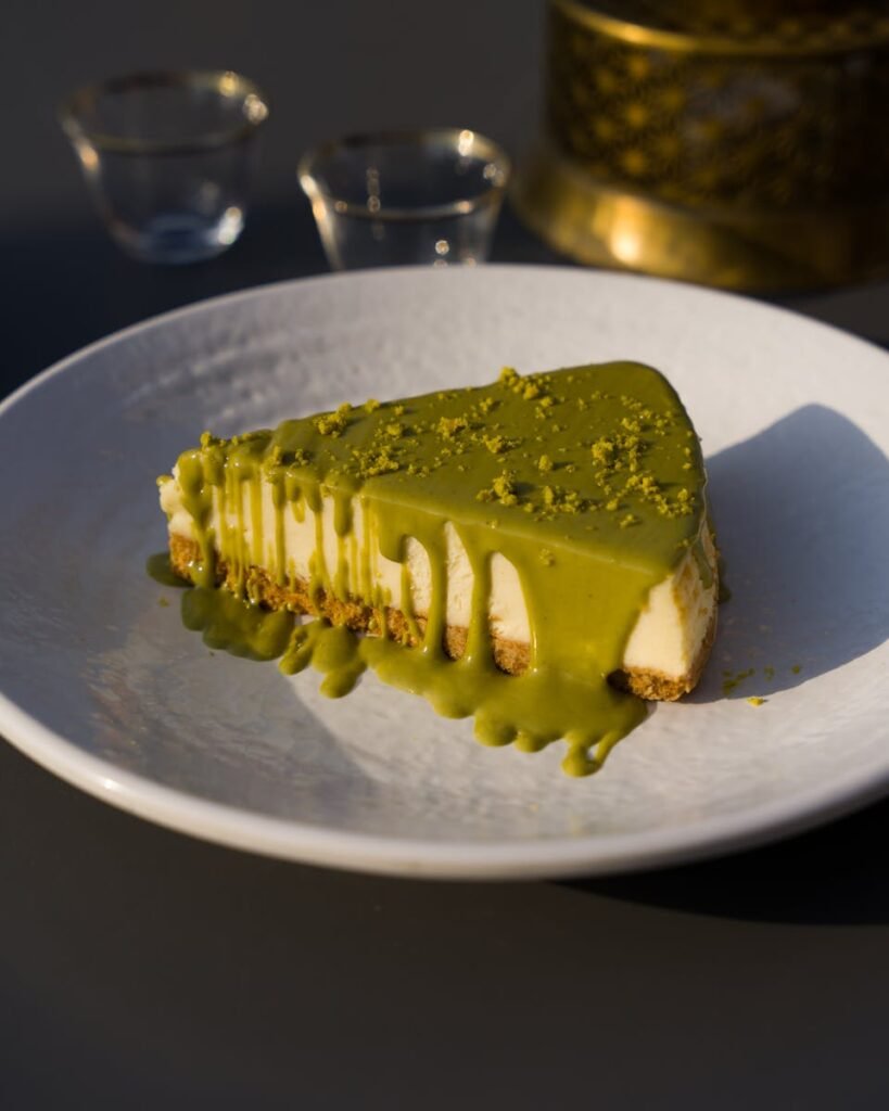 A close-up of a creamy cheesecake slice drizzled with rich matcha glaze on a white plate.